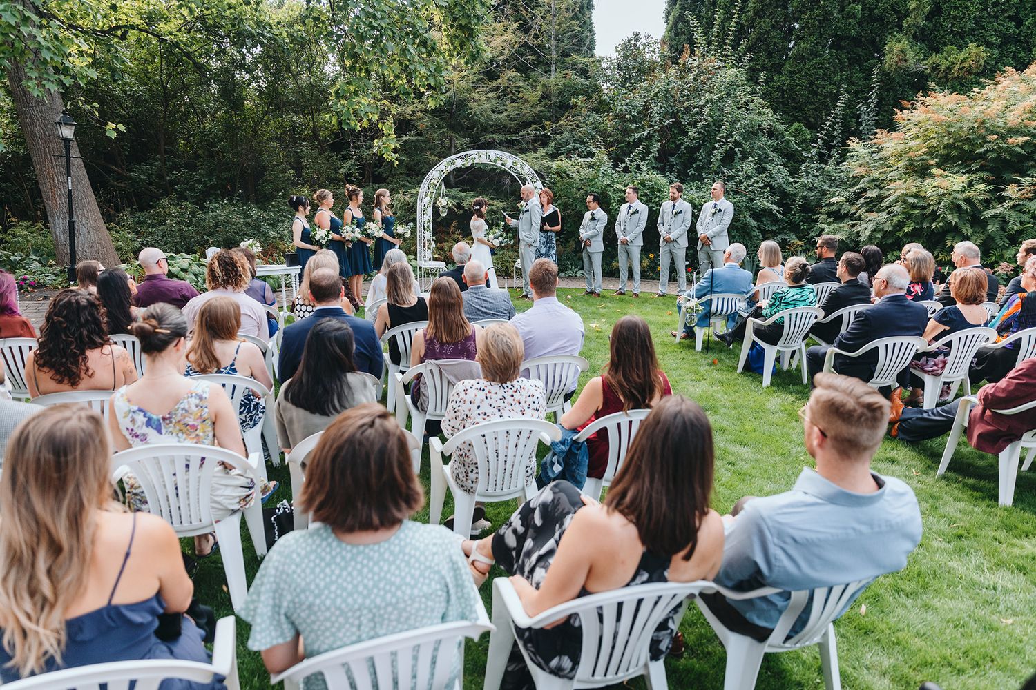 Wedding guests seated in white chairs on a lush green lawn during an outdoor garden ceremony.