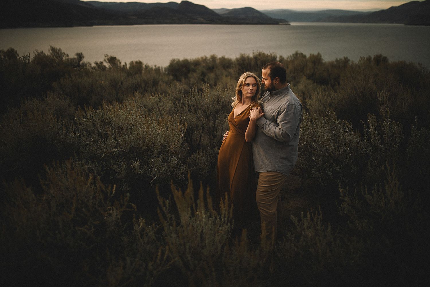 Silhouetted figures embrace during golden hour in a rustic field with mountains and lake in the background.