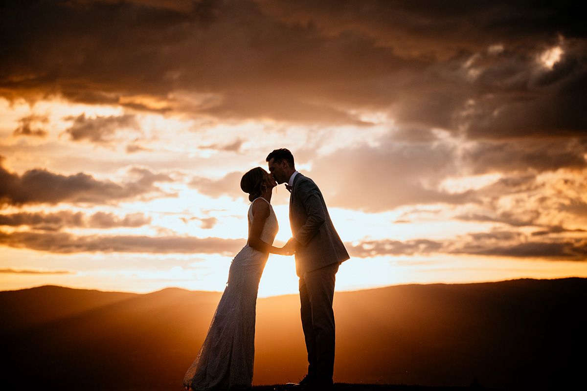 Silhouettes of a couple share a romantic moment at sunset with dramatic orange sky and mountain backdrop.