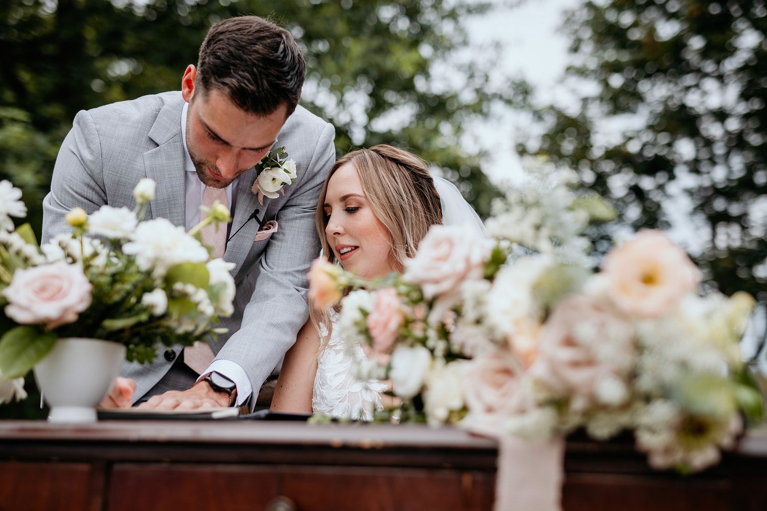 A bride and groom sign marriage certificate in soft natural light with delicate pink floral decorations in the background.