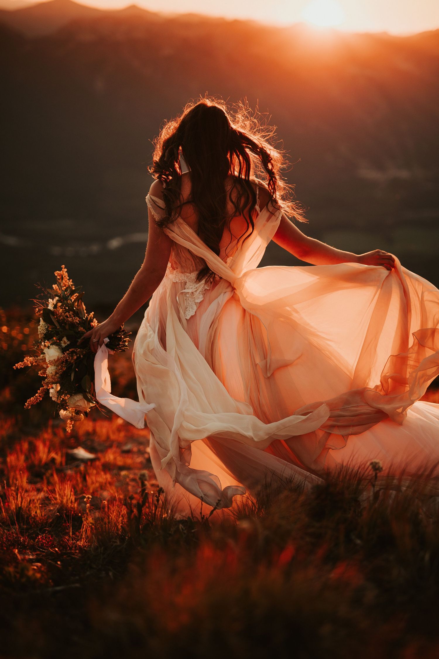 A bride in a flowing white dress dances gracefully in a field at sunset, creating a dreamy silhouette against the orange sky.
