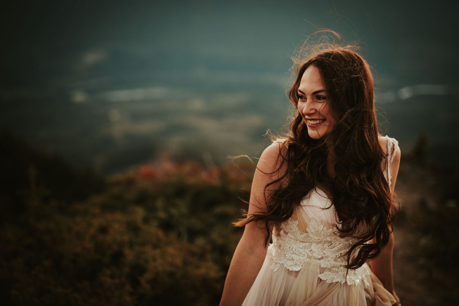 A bride in a white dress smiles while standing on a hilltop during golden hour with a blurred landscape background.