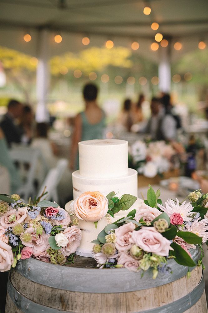 Two-tier white wedding cake decorated with blush pink roses and greenery sits atop rustic wine barrel under string lights.