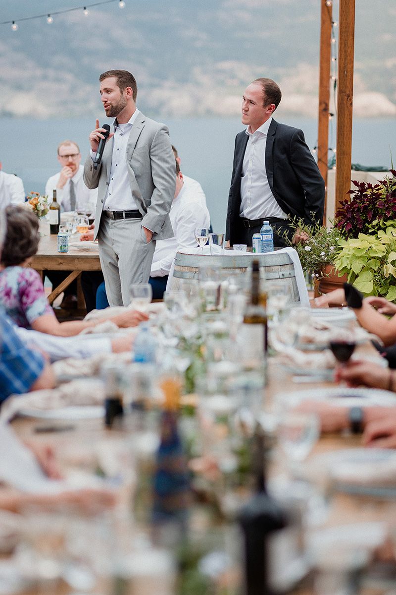 Vows and Vines Vineyard wedding guests seated at outdoor reception tables with string lights overhead and blurred glassware in foreground.