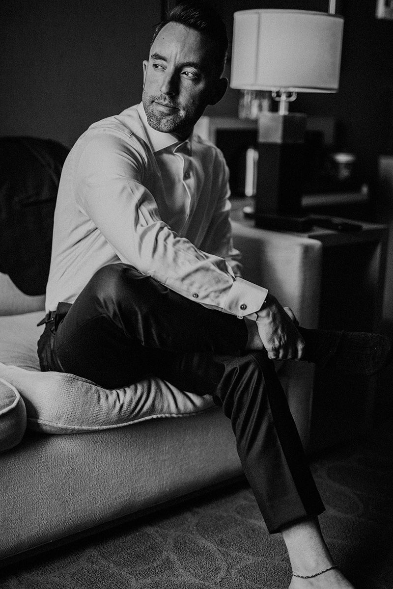 Black and white portrait of a man seated on a couch wearing a light colored shirt and dark pants in a moody setting.