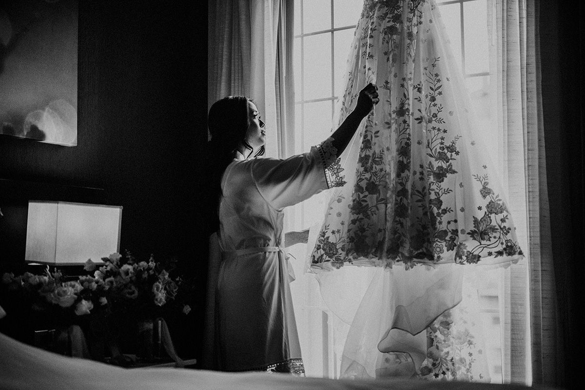 A silhouetted bride in her morning robe, adjusts her elegant lace wedding dress by a bright window in a black and white photograph.