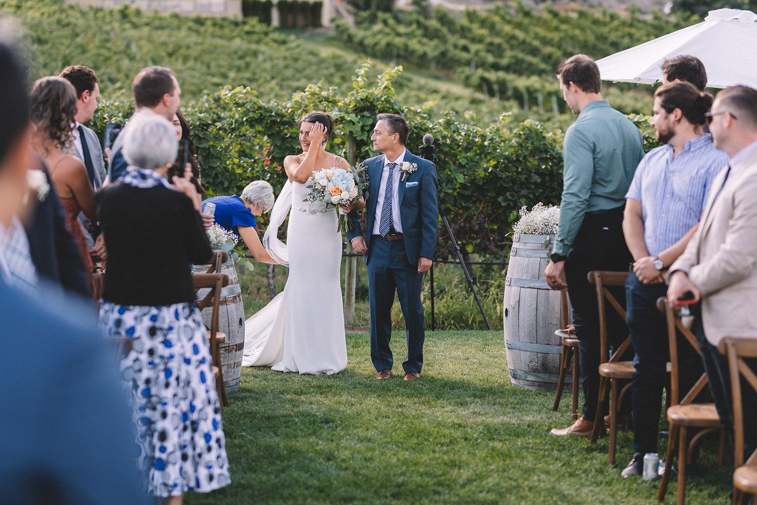 Wedding guests gather outdoors at a vineyard ceremony with rows of grapevines and rolling vineyards in the background.