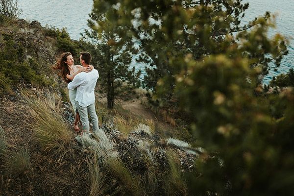 Couple embracing on a rocky hillside surrounded by pine trees with a scenic lake view in the background.
