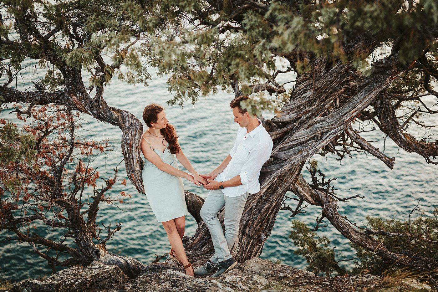 Couple holding hands among twisted tree branches by turquoise water, romantic outdoor photo session.