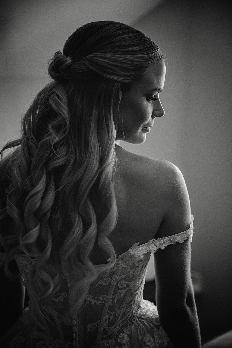 Black and white portrait of a bride in an off-shoulder lace dress with long wavy hair styled in a half-up braided design.