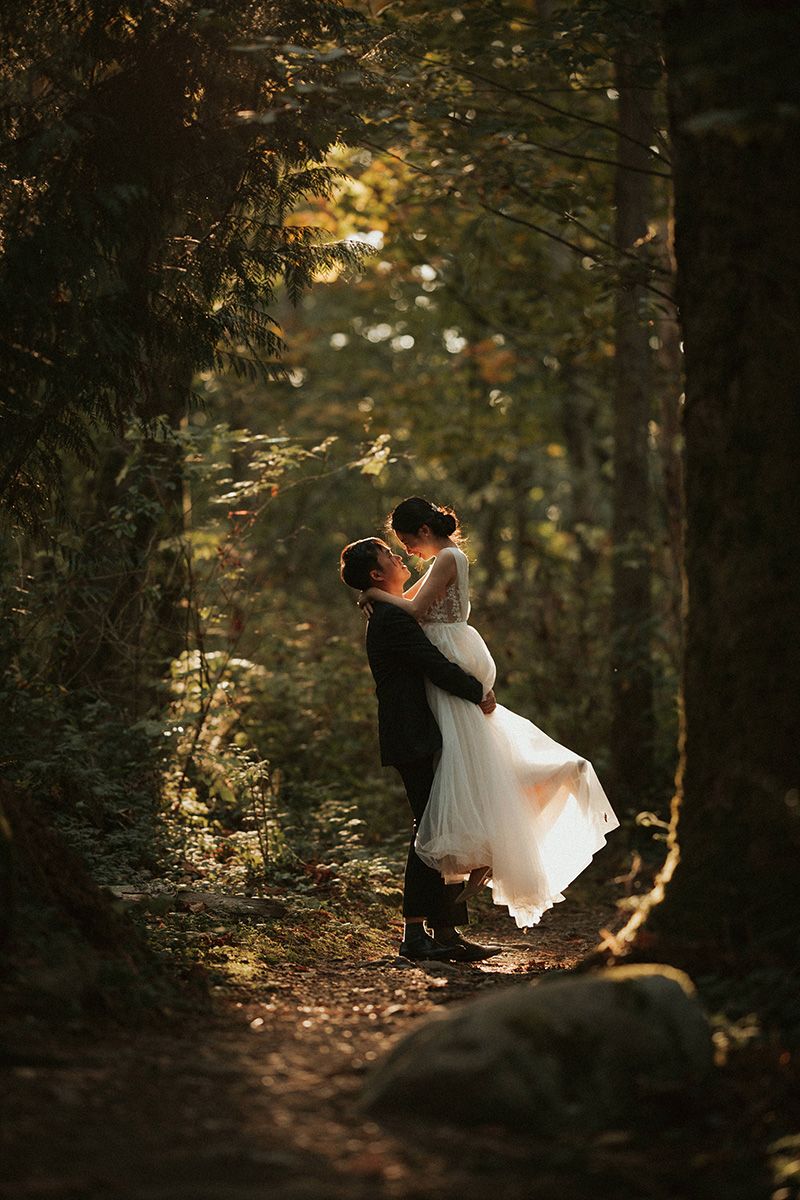 A groom lifts his bride off the ground on a sunlit forest path with golden light filtering through the trees.