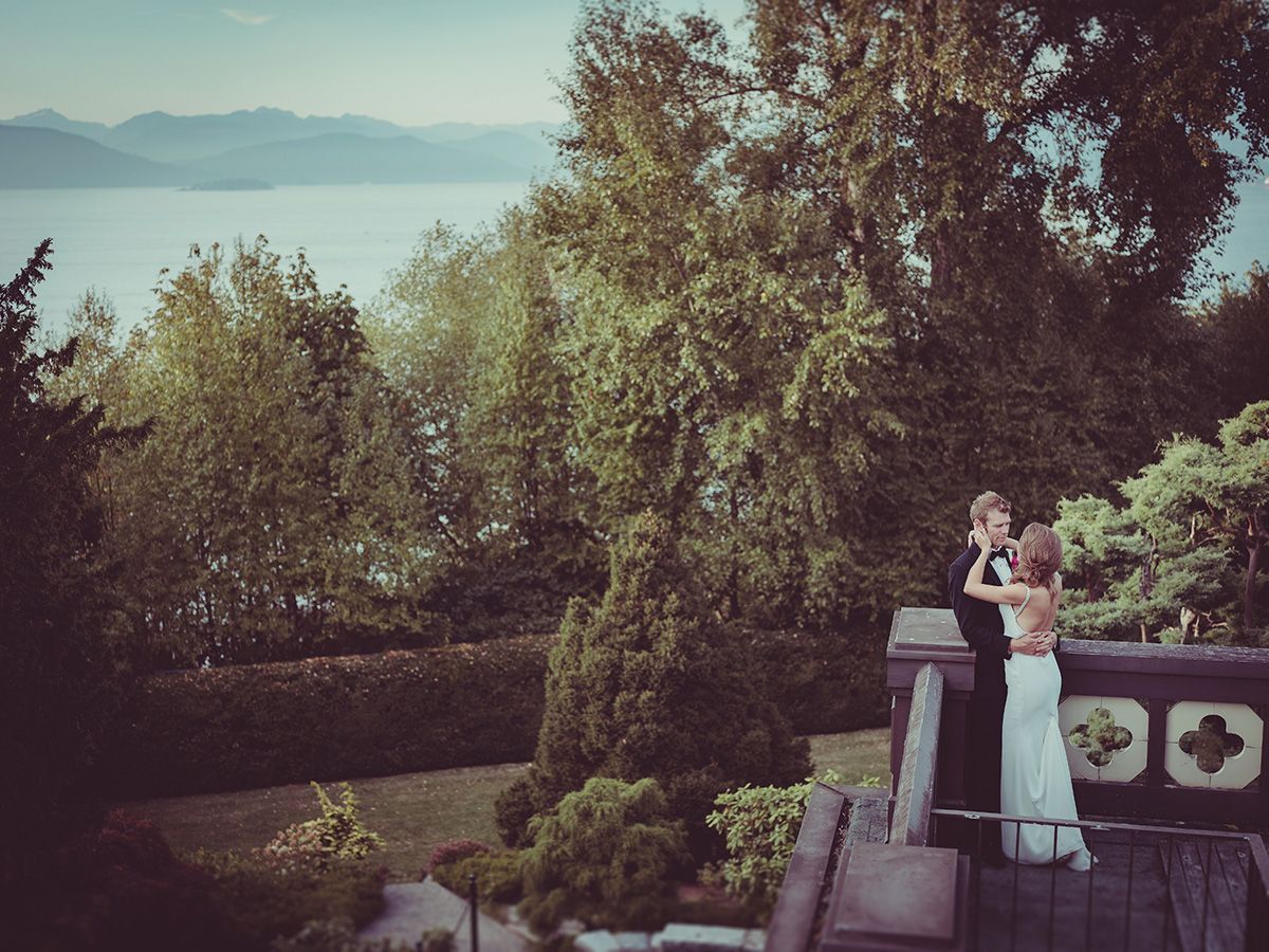 Bride and groom cuddle together on the balcony of Cecil green Park House in Vancouver with gardens, ocean and mountains in the background.