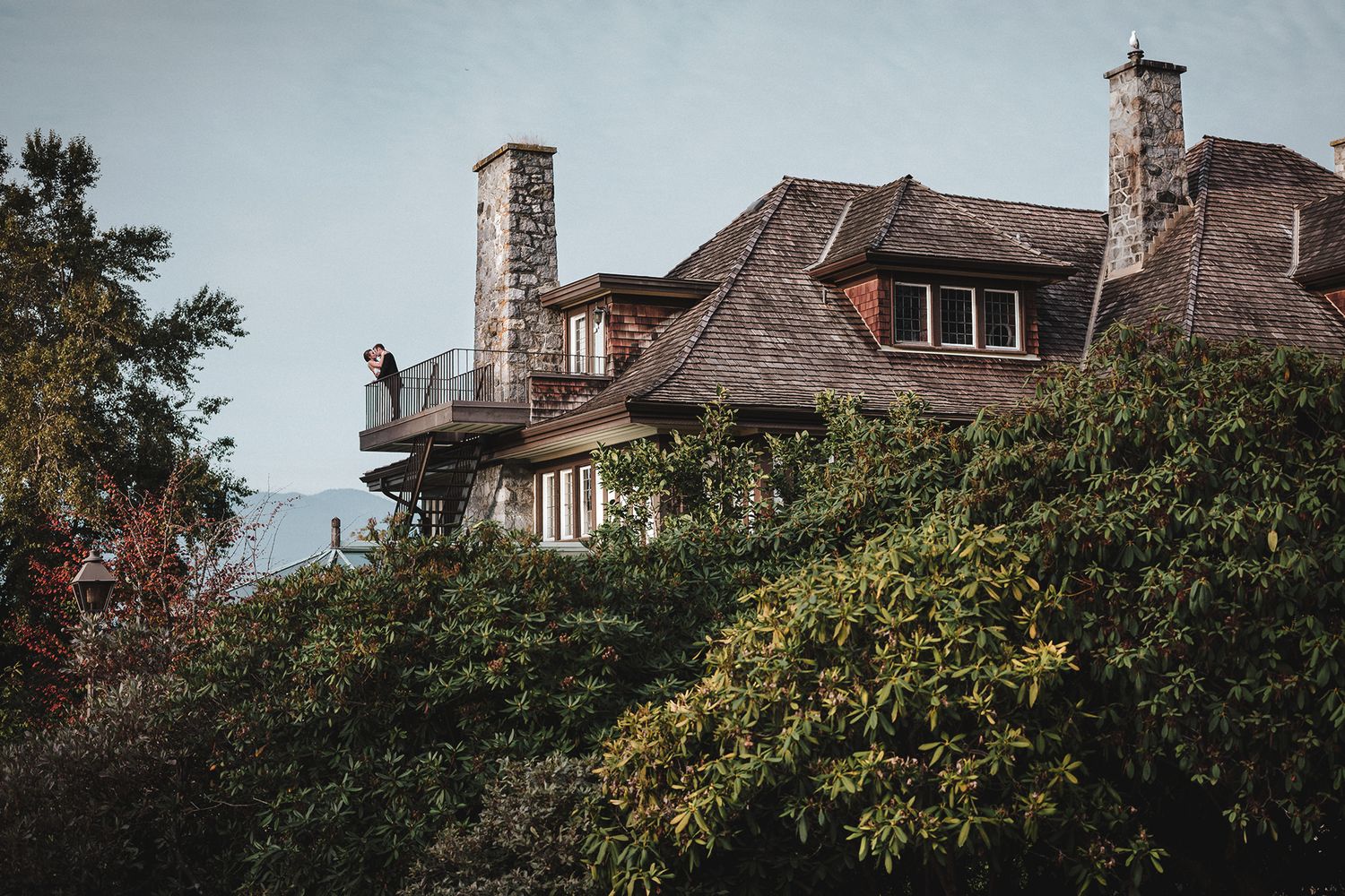 Stone mansion with turret and dormer windows sits among lush trees against cloudy sky. A man kisses a woman on the balcony.