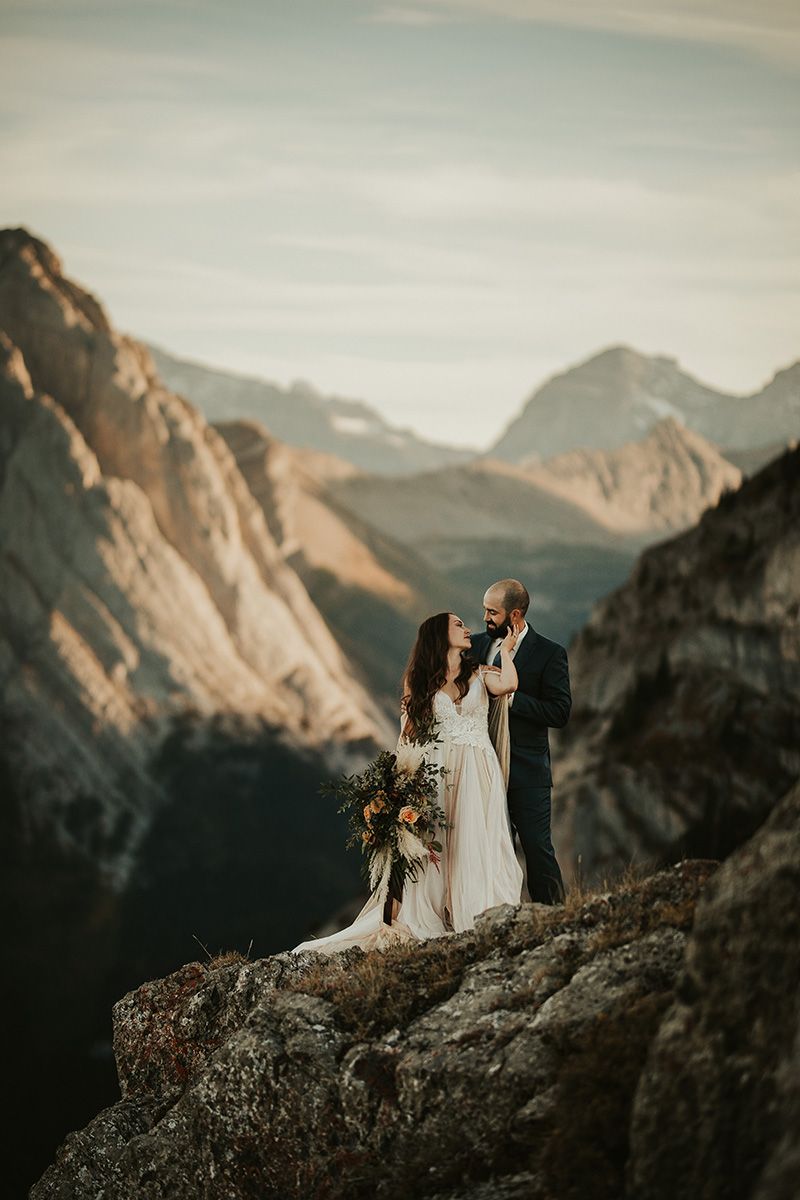 A couple in wedding attire stands on a mountain peak overlooking dramatic mountain ranges at sunset among the Canadian Rockies.