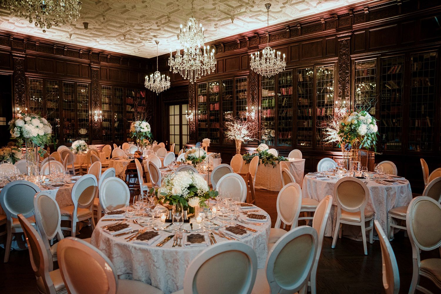 Elegant wedding reception hall with crystal chandeliers and round tables set with white linens and floral centrepieces.