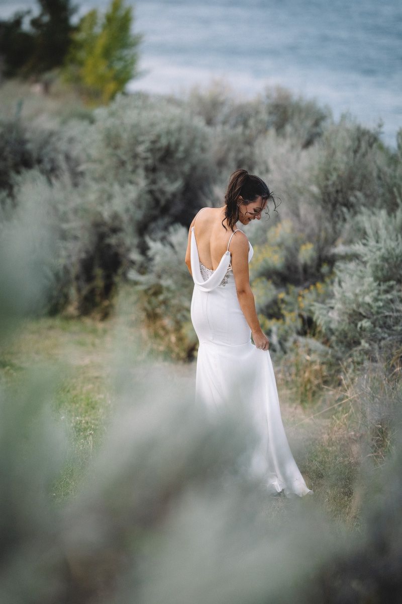 A bride in a flowing white dress walks through sage brush and desert vegetation against a soft blurred background.