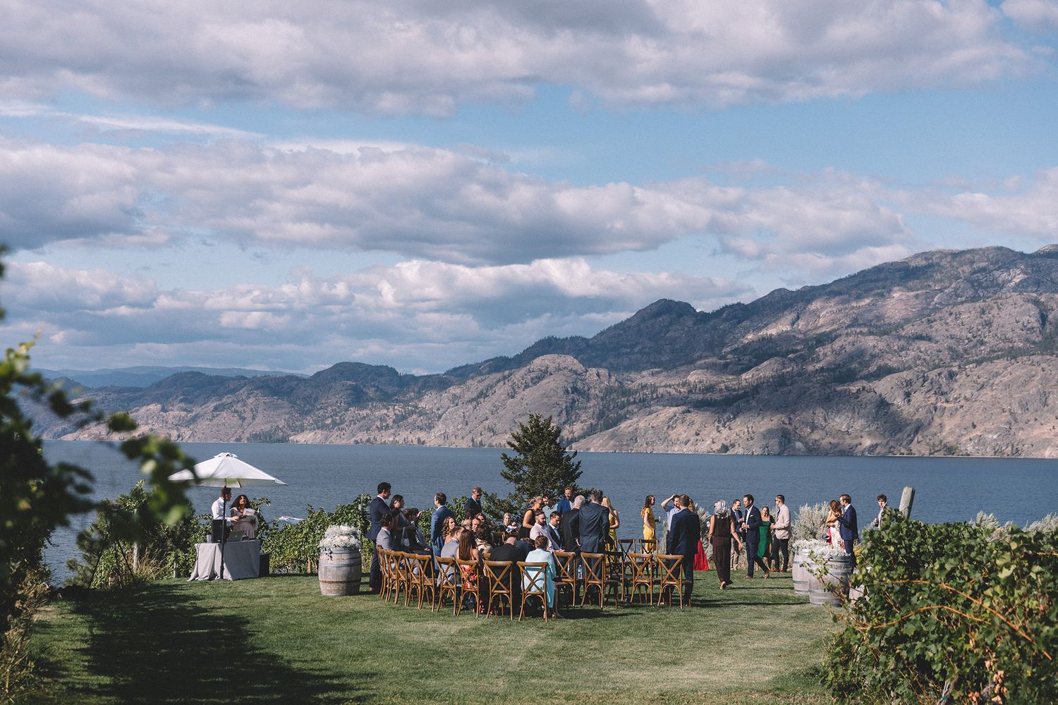 A wide shot of a wedding ceremony on a lawn surrounded by vineyards with lake Okanagan in the background.