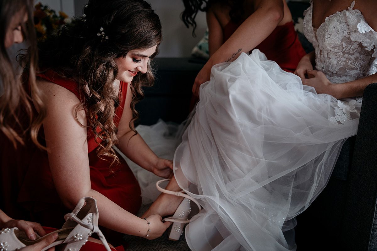 A bride in a white wedding dress is being helped with final preparations by attendants in red dresses.