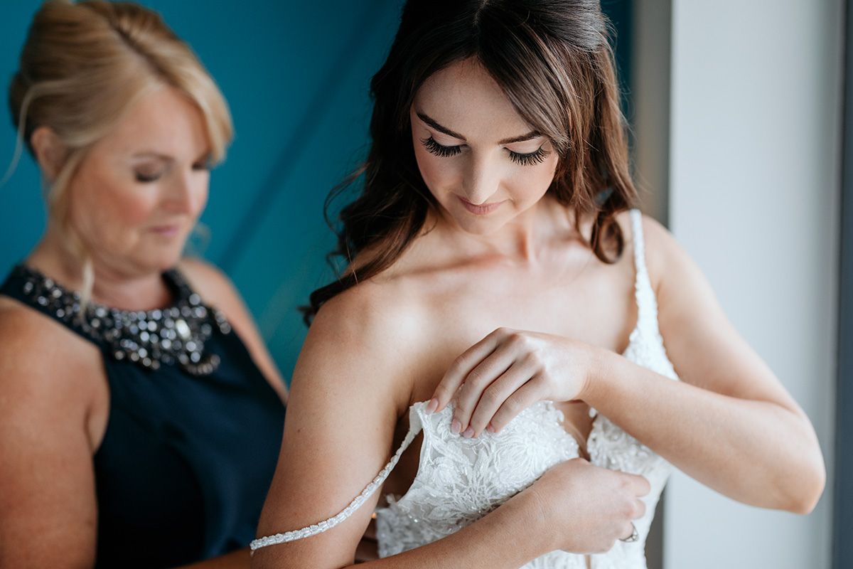 Wedding party members help adjust a bride's dress and accessories against a teal colored background.