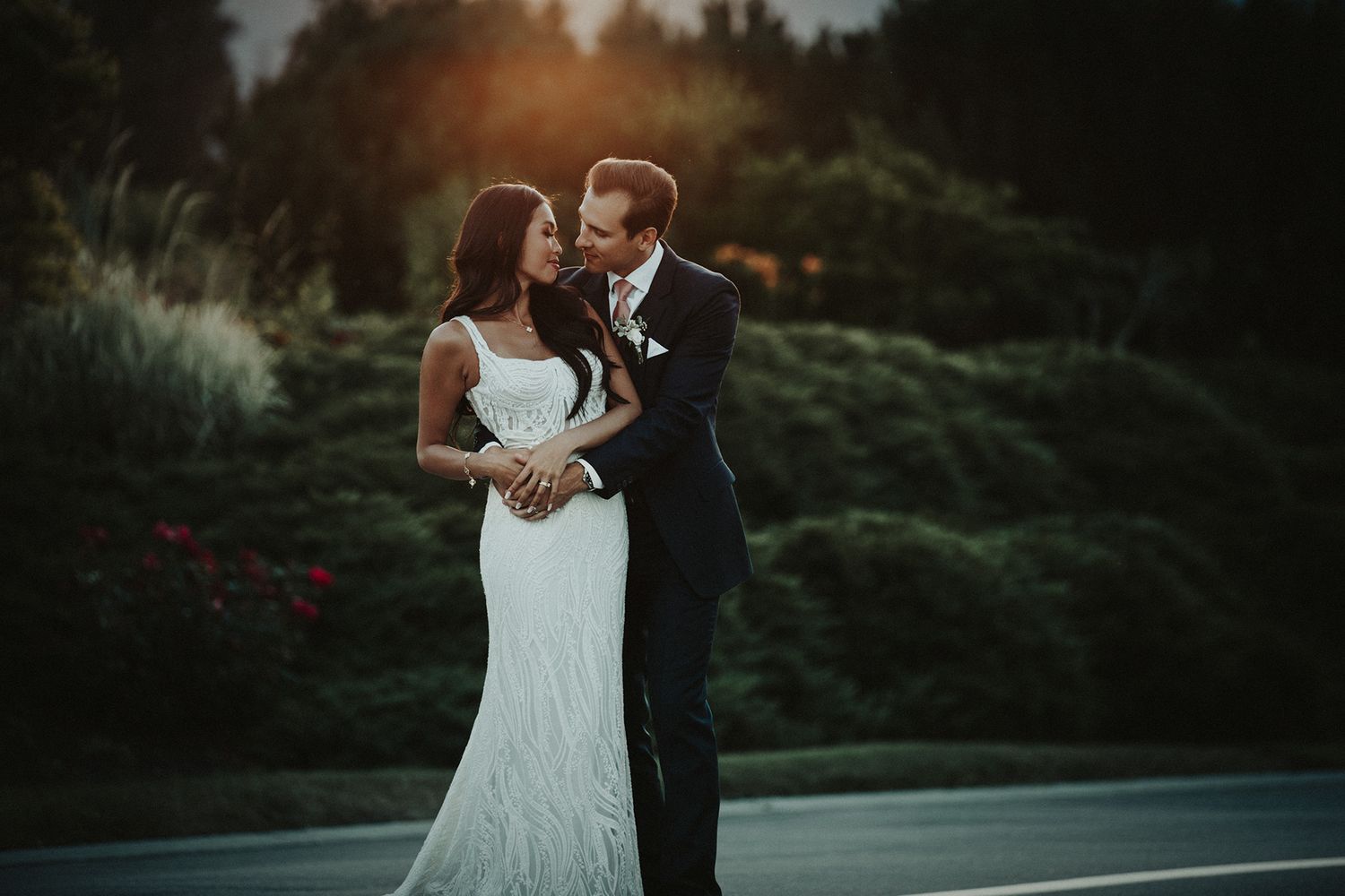 A couple in wedding attire embraces during golden hour with lush greenery and flowers in the background at The Harvest Golf Club.