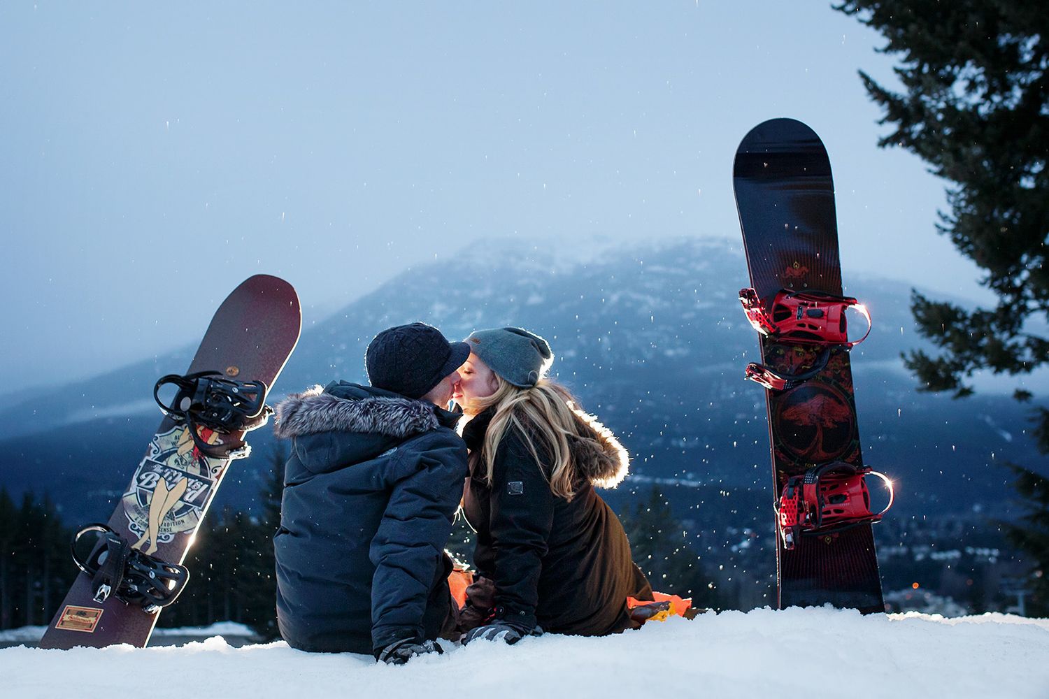 Snowboarder couple share a kiss at the summit of a snowy mountain with scenic mountain ranges in the background.