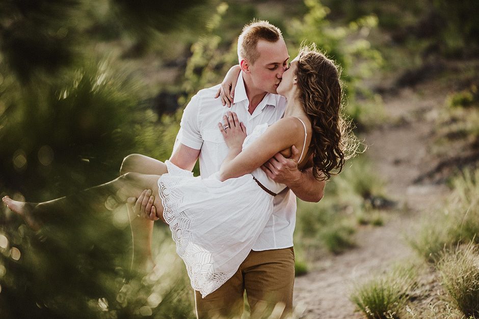 A man carries his girlfriend in his arms as he walks a mountain path at sunset.