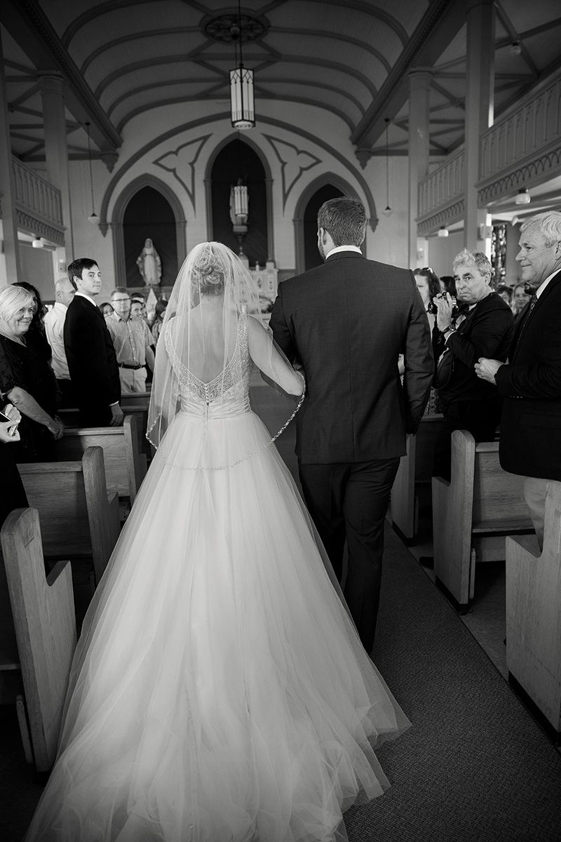 A black and white wedding photo showing a bride in a flowing ballgown walking down the church aisle.