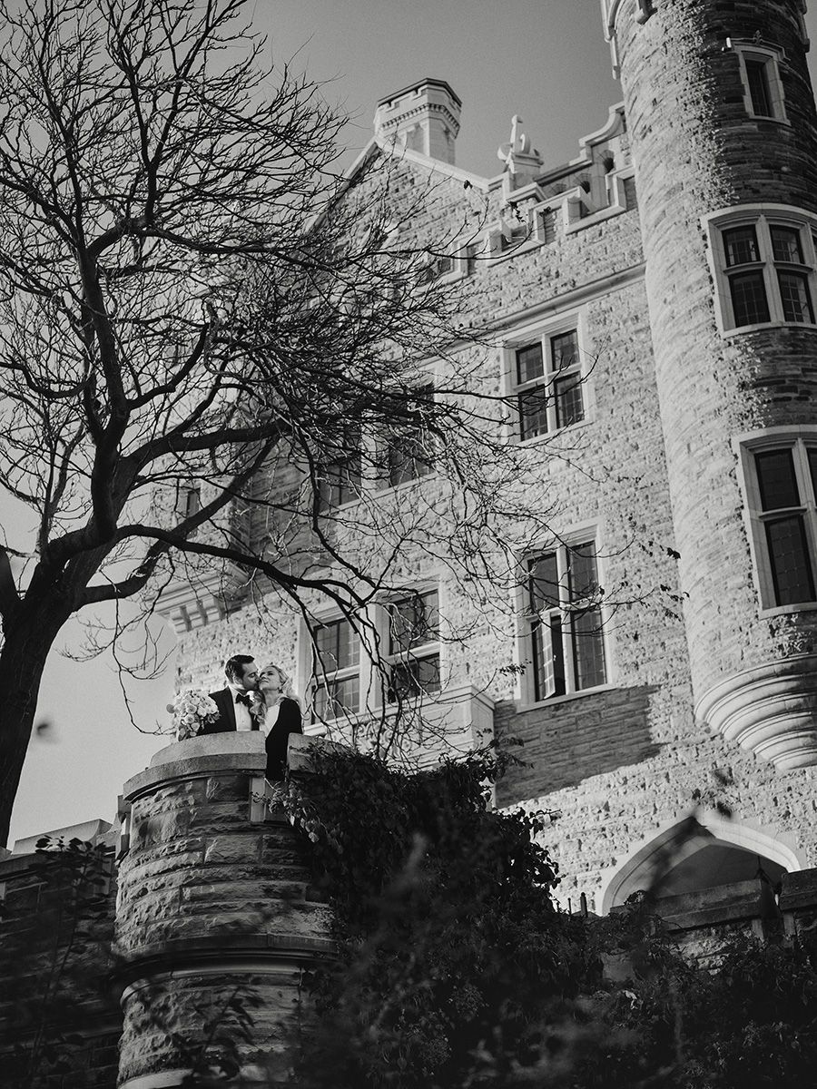 A black and white architectural photograph shows a stone castle-like building with turrets and bare winter trees against the sky.