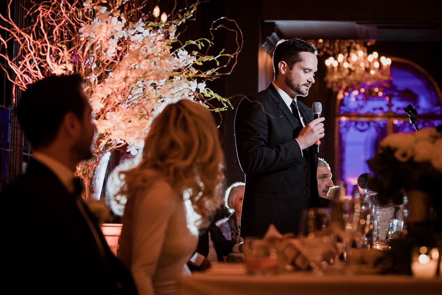 Bride looks on as best man gives speech at elegant wedding reception with candlelit tables and crystal chandeliers.