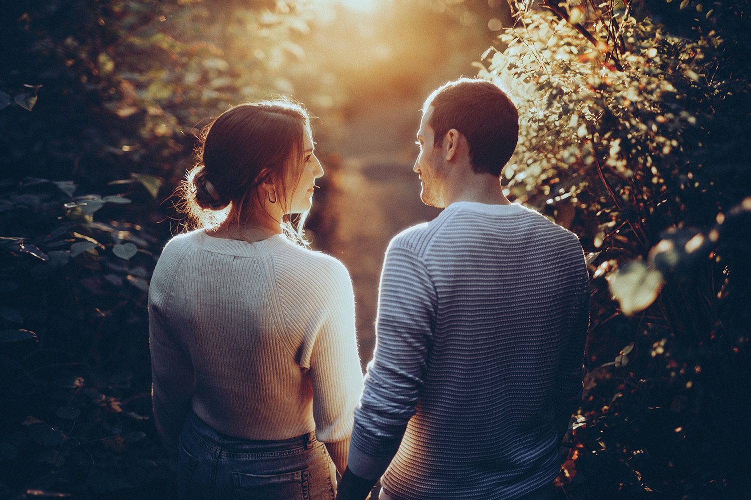 Couple walking together in a sunlit forest path during golden hour as sunlight filters through the trees.