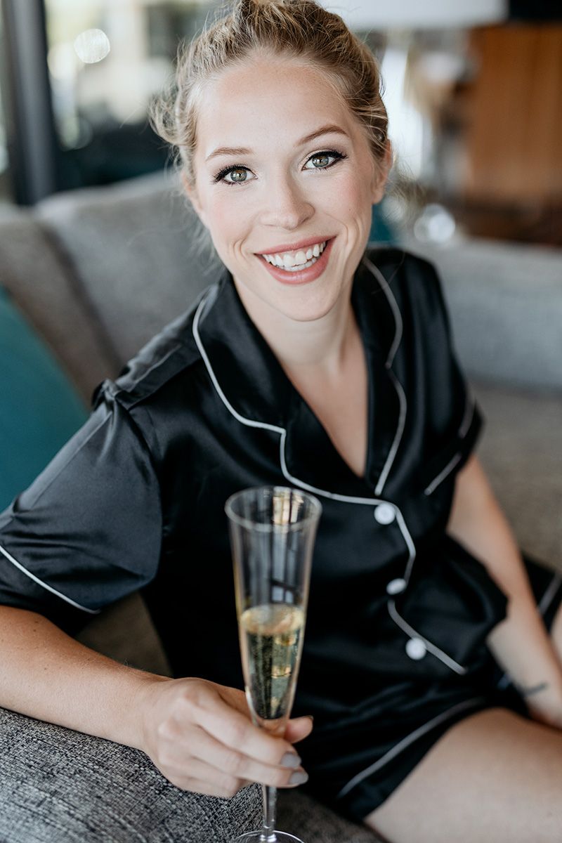 A woman in black silk pajamas holding champagne flute while relaxing on gray couch with bright smile.