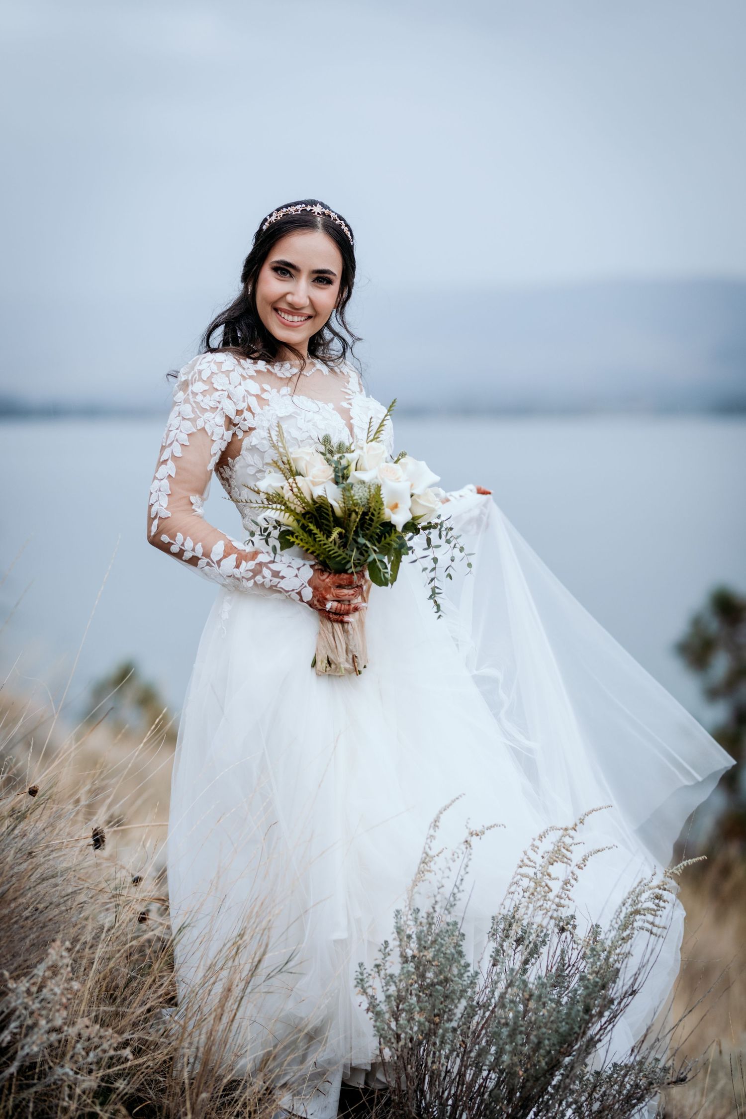 A bride in a white lace dress stands among tall grass holding a bouquet against a misty mountain and lake backdrop.