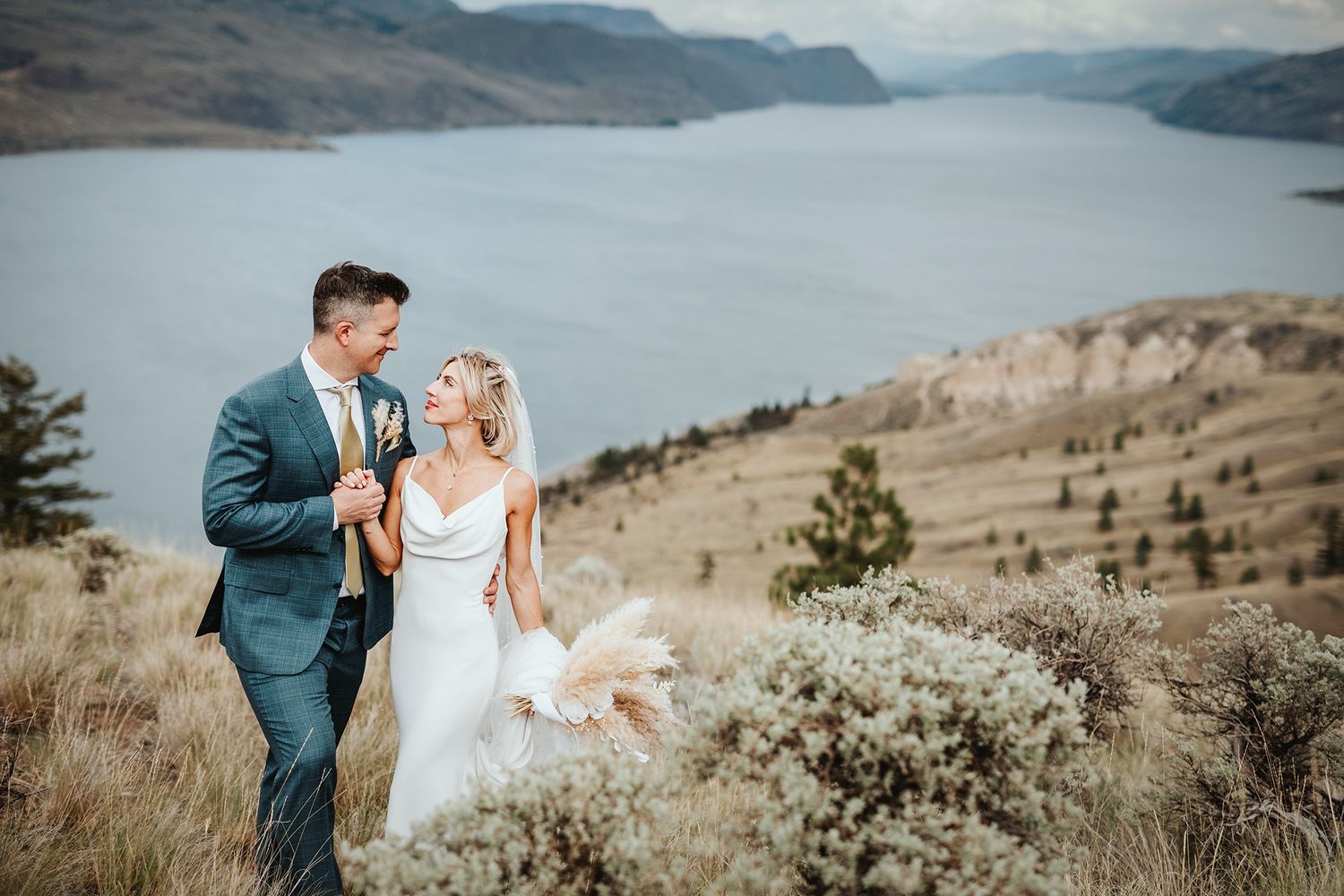 A couple in wedding attire stands on a scenic mountain overlook with a lake and rolling hills in the background.