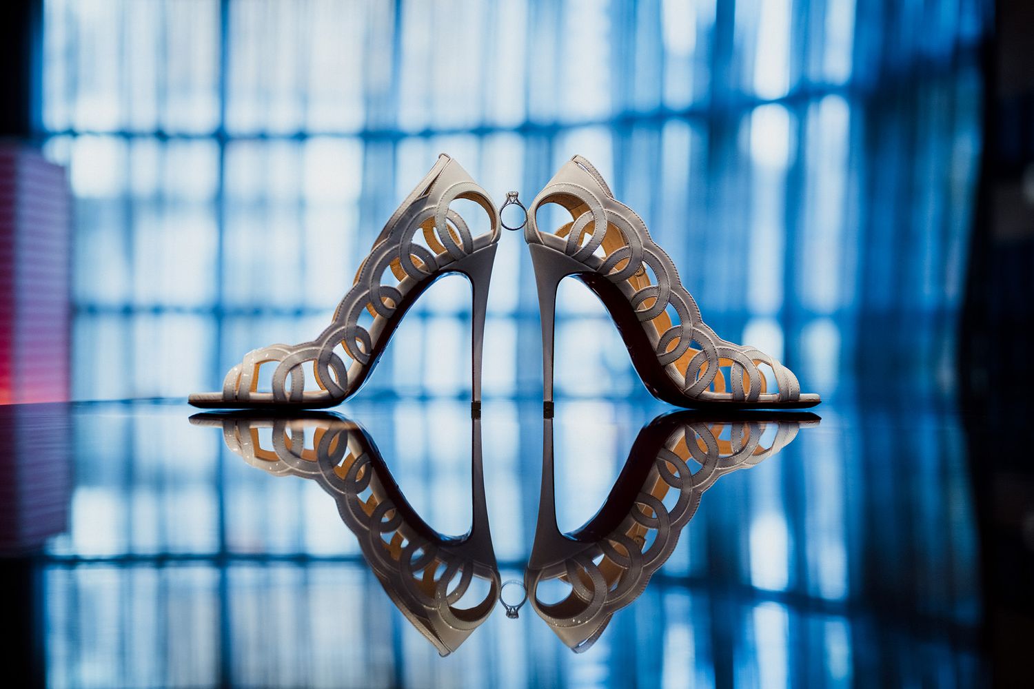 An artistic photo of wedding shoes sitting on a reflective surface with an engagement ring sitting between them.