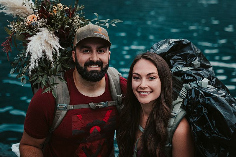 A couple poses together near a turquoise lake with mountains in the background during a hiking adventure.