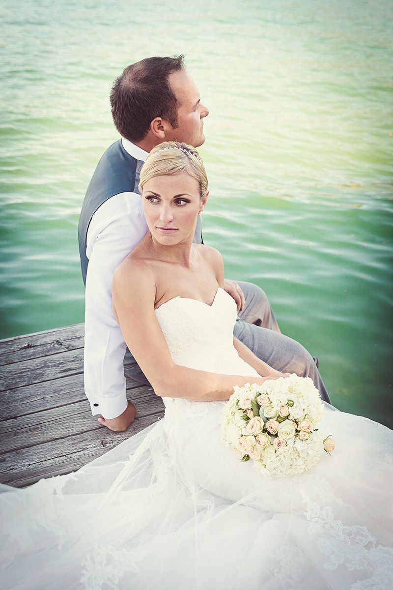 A romantic wedding couple sits on a dock by turquoise water with the bride holding a soft cream-colored bouquet.