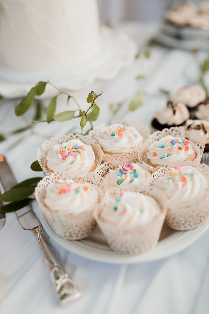 White frosted cupcakes with colourful sprinkles displayed on delicate lace doilies and white plates with greenery accents.