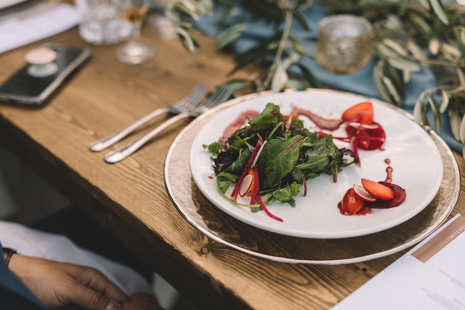 Fresh strawberries and mixed greens arranged on white plates with elegant table settings on a wooden table.