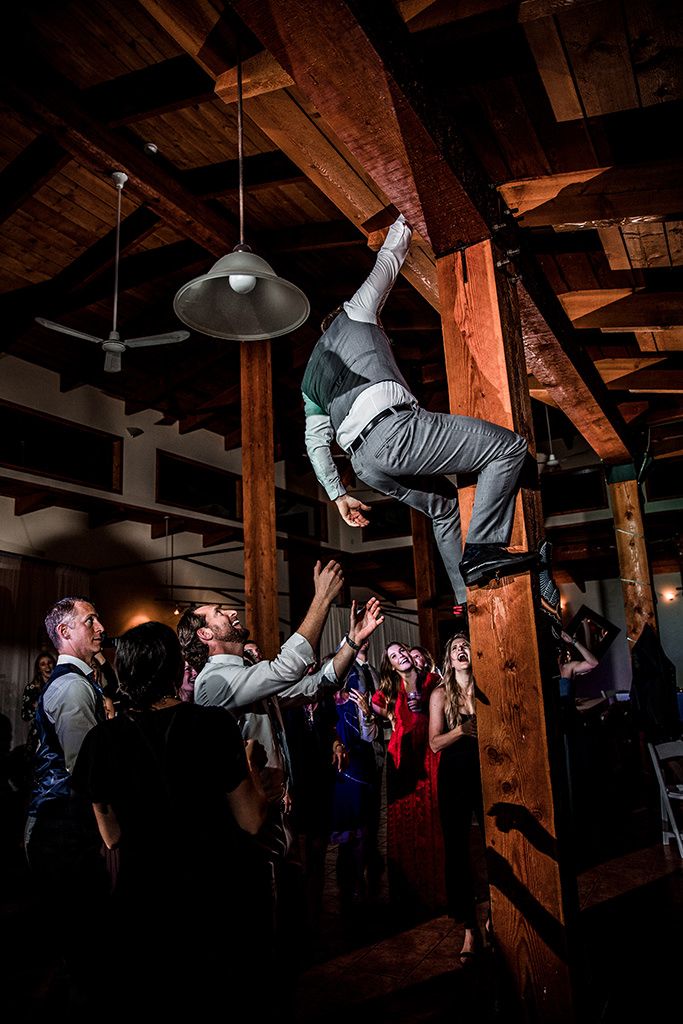 A person climbs a wooden beam to retrieve a bride's garter at an indoor venue with exposed wooden beams while party-goers watch from below.