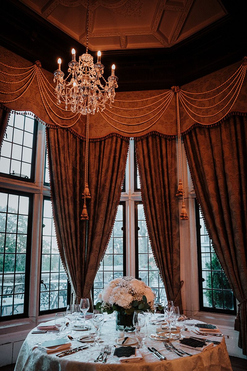 Elegant wedding reception tables set with crystal chandeliers and dramatic burgundy drapes in a dim banquet room.