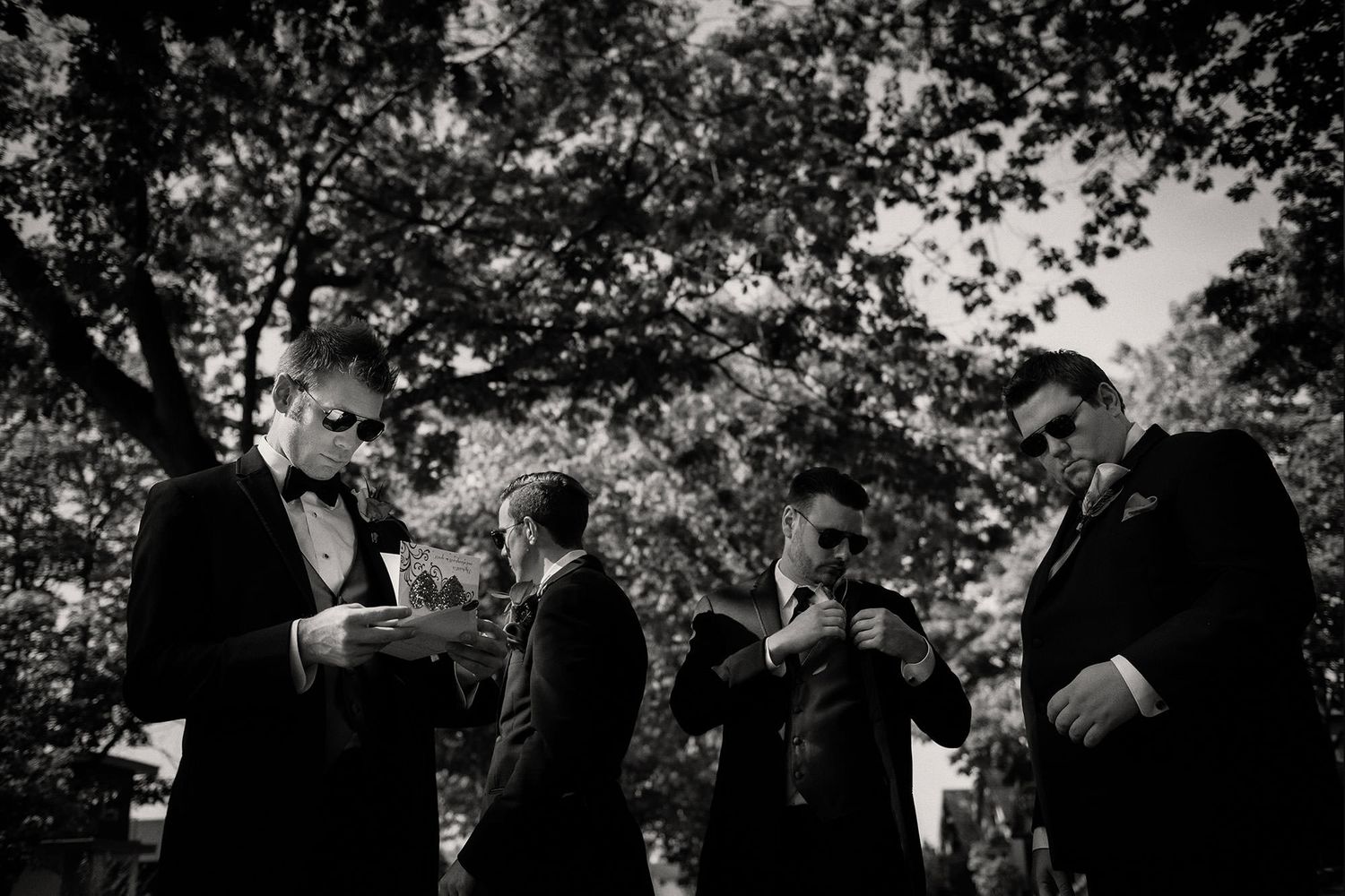Black and white candid photograph of people gathered outdoors under tree branches in formal attire.