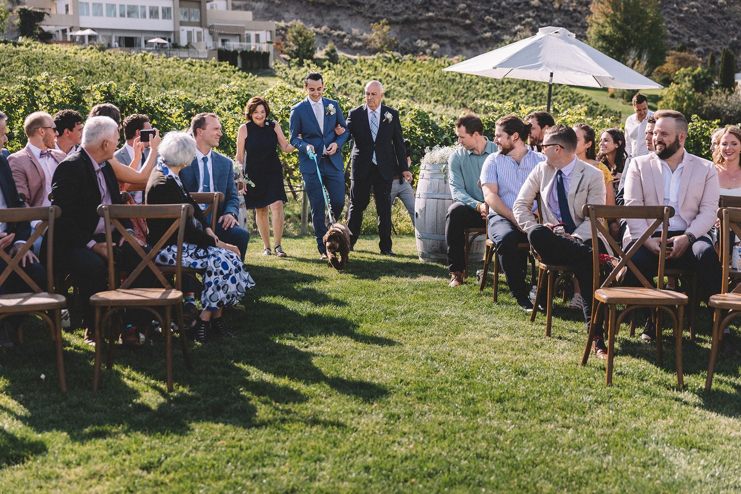 Wedding ceremony setup on a grassy hillside with wooden chairs and guests gathered under sunny mountain scenery.