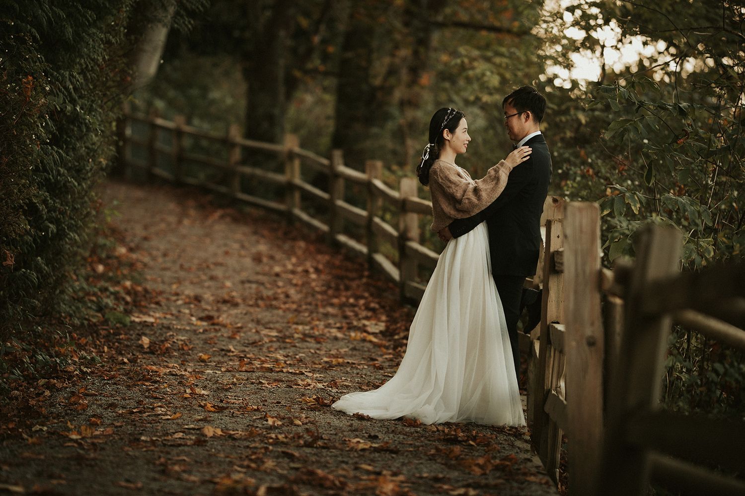 A romantic couple in wedding attire embrace on a rustic autumn path lined with wooden fencing and fallen leaves.