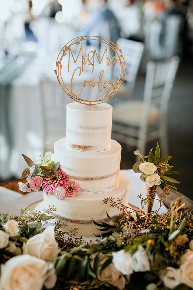 Elegant three-tier white wedding cake decorated with roses and greenery, topped with a gold 'Mr & Mrs' cake topper. Summerhill Winery.