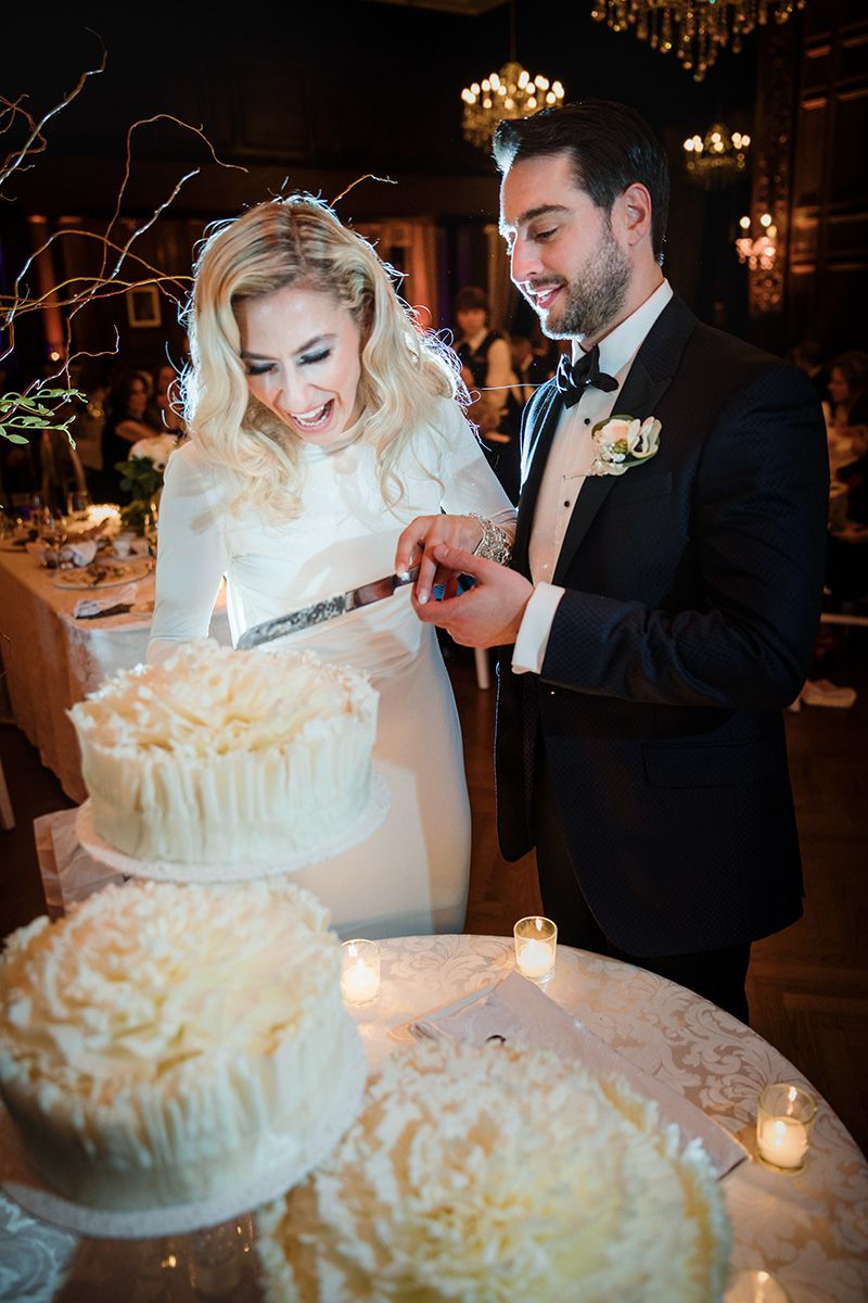 A couple cuts their elegant white wedding cake with multiple tiers in a dimly lit reception venue with chandeliers.