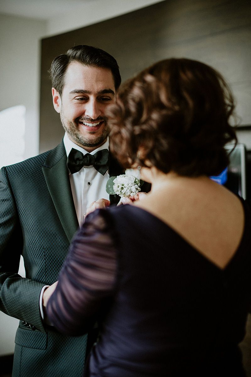 A tender moment at a formal event as a woman pins a white boutonniere onto a black tuxedo jacket.