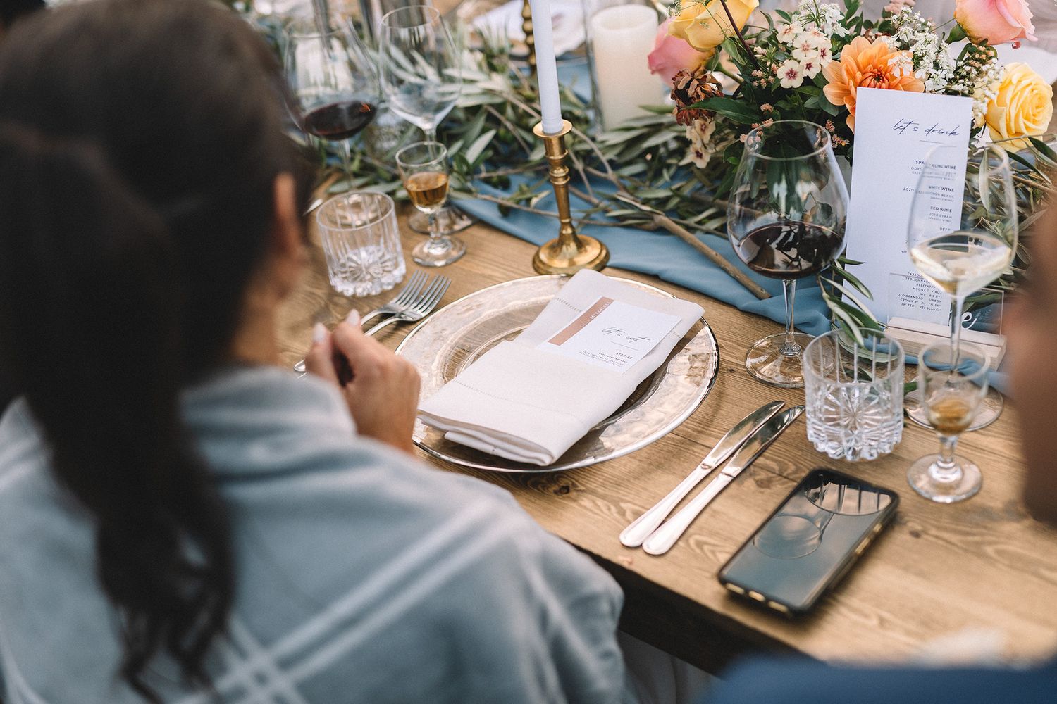 People gathered around a rustic wooden table at a wedding reception with elegant place settings and floral decor.