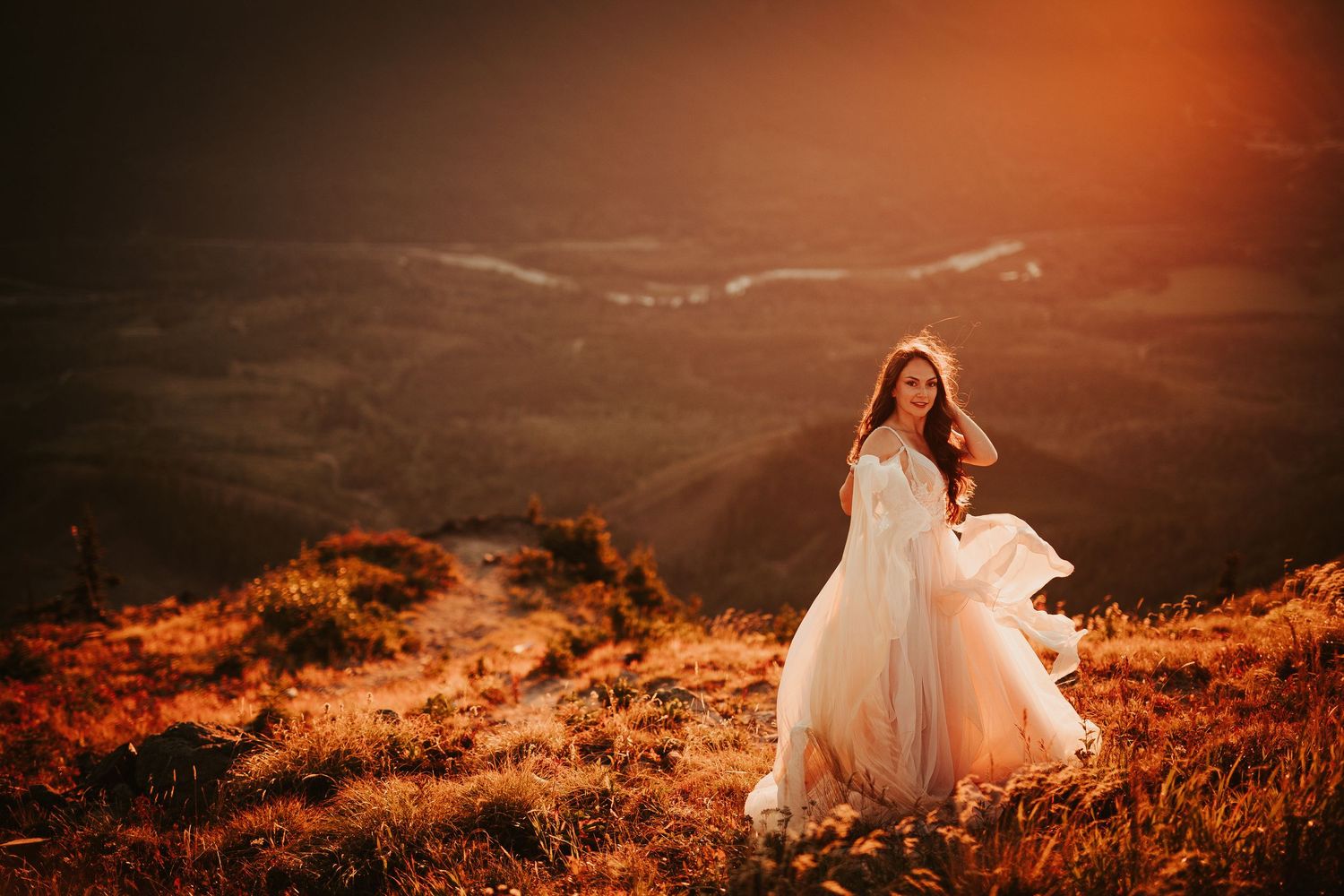 A bride in a flowing white dress stands on a hilltop against a dramatic sunset sky.