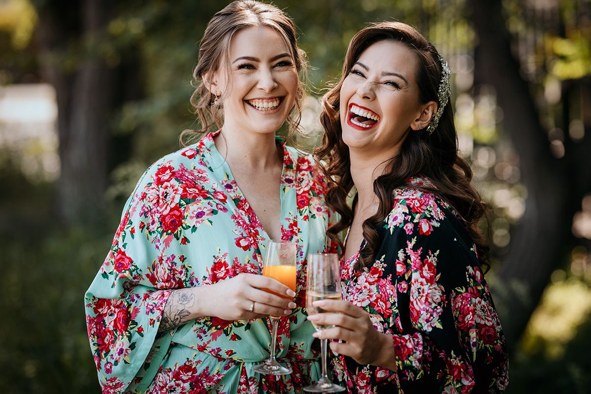 Two friends wearing floral robes laugh together while holding drinks during an outdoor celebration.