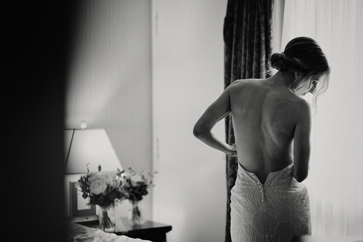 Black and white photo showing intimate moments of a bride's bare back as she is getting dressed in silhouette against window light.